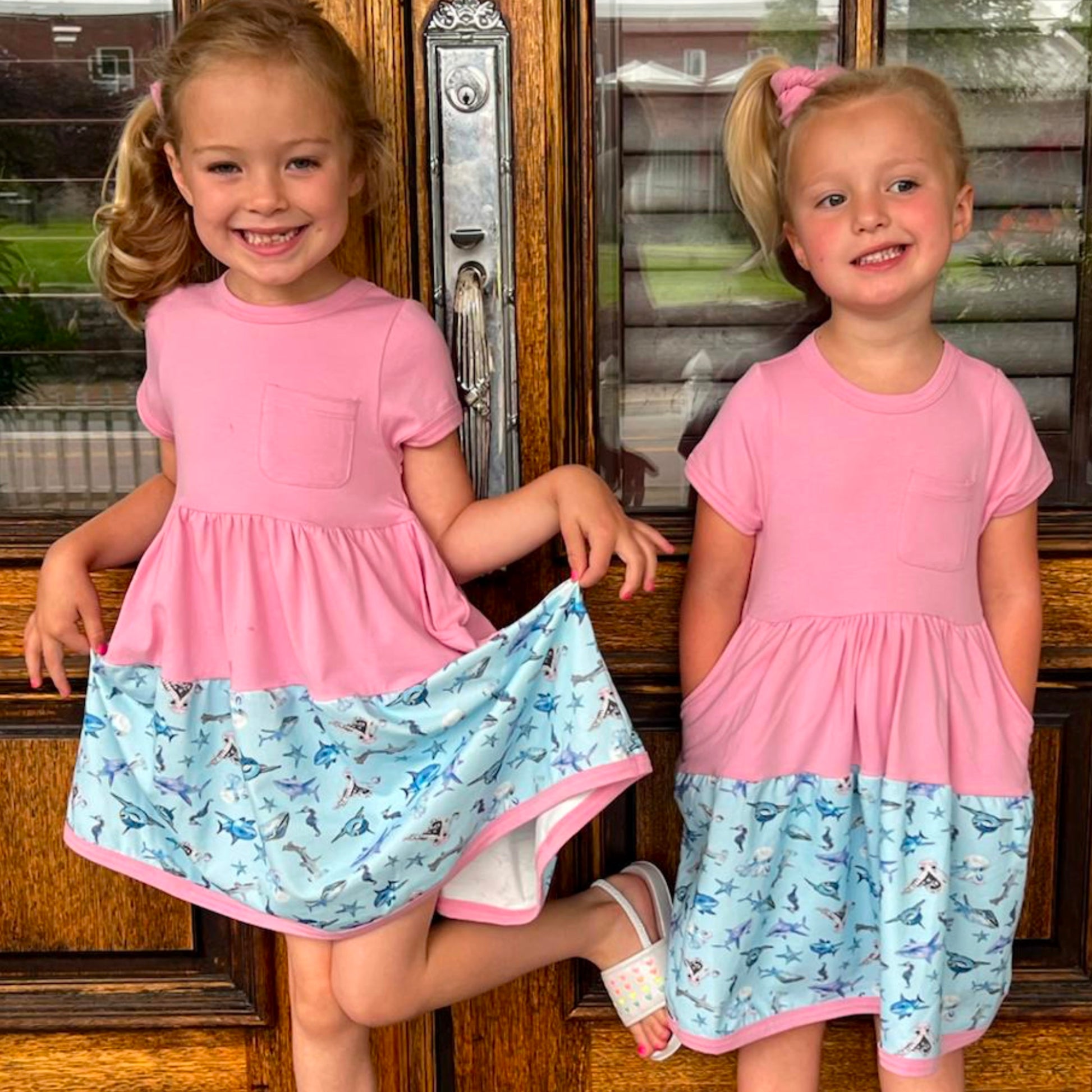 Two girls wearing pink and blue Sparky Shark dresses – short-sleeve twirl dresses with matching scrunchies and sea creature print with matching scrunchies