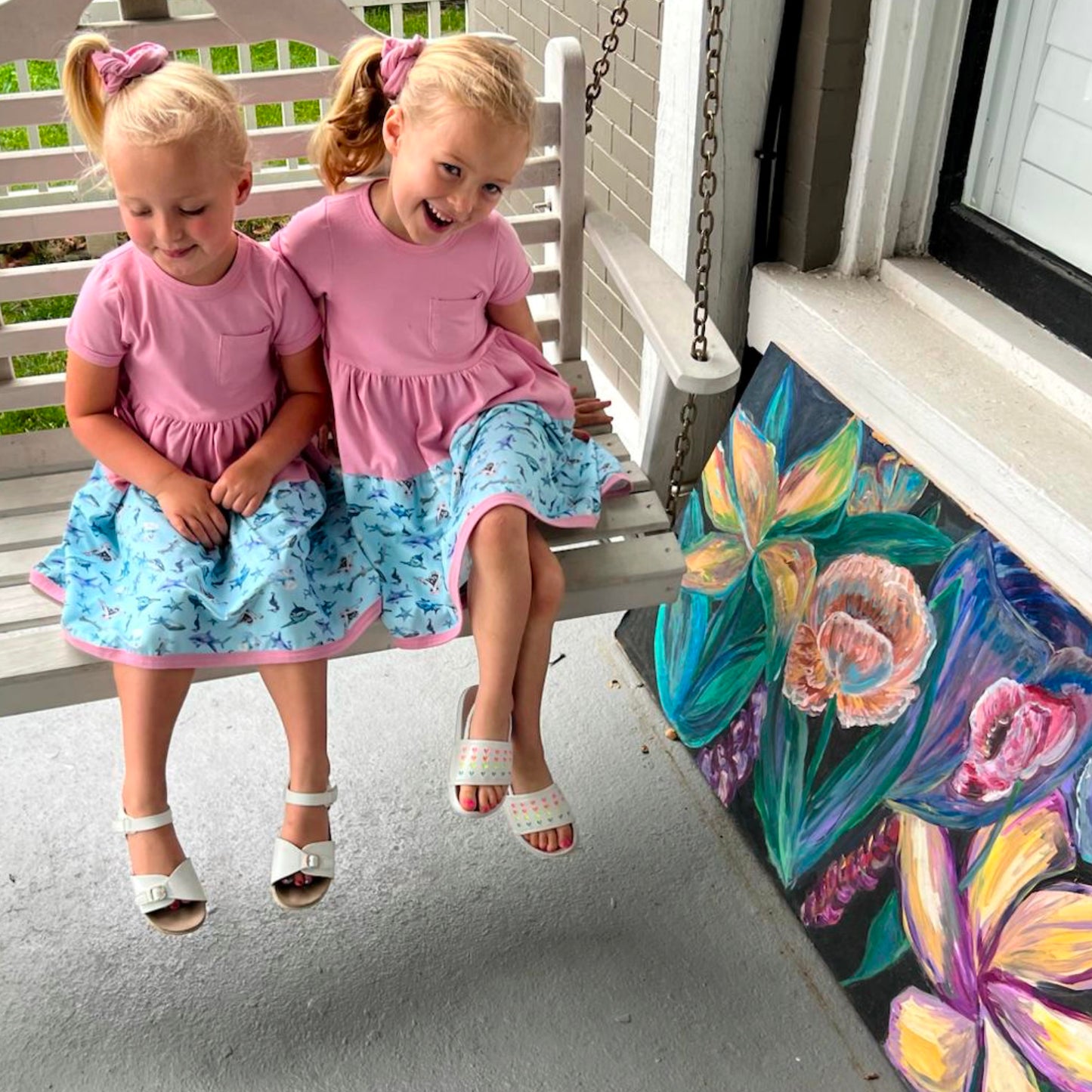 Two girls wearing pink and blue Sparky Shark dresses – short-sleeve twirl dresses with matching scrunchies and sea creature print