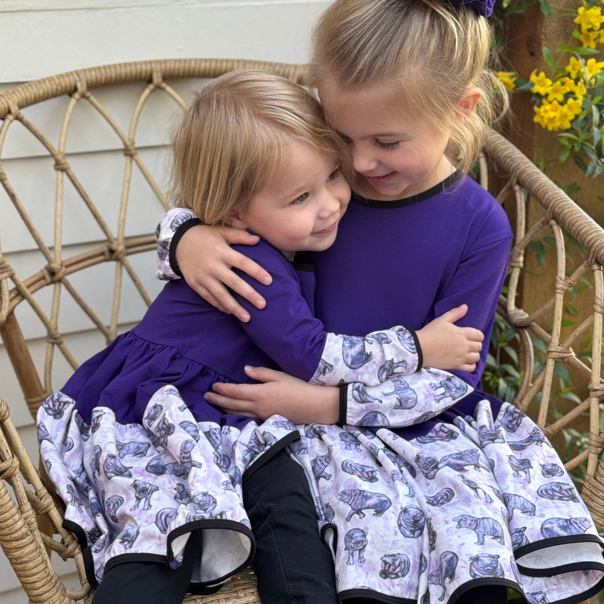 Two young girls wearing matching purple hippo dresses, smiling and playing together outdoors. The dresses feature a fun animal print and twirl-friendly design.